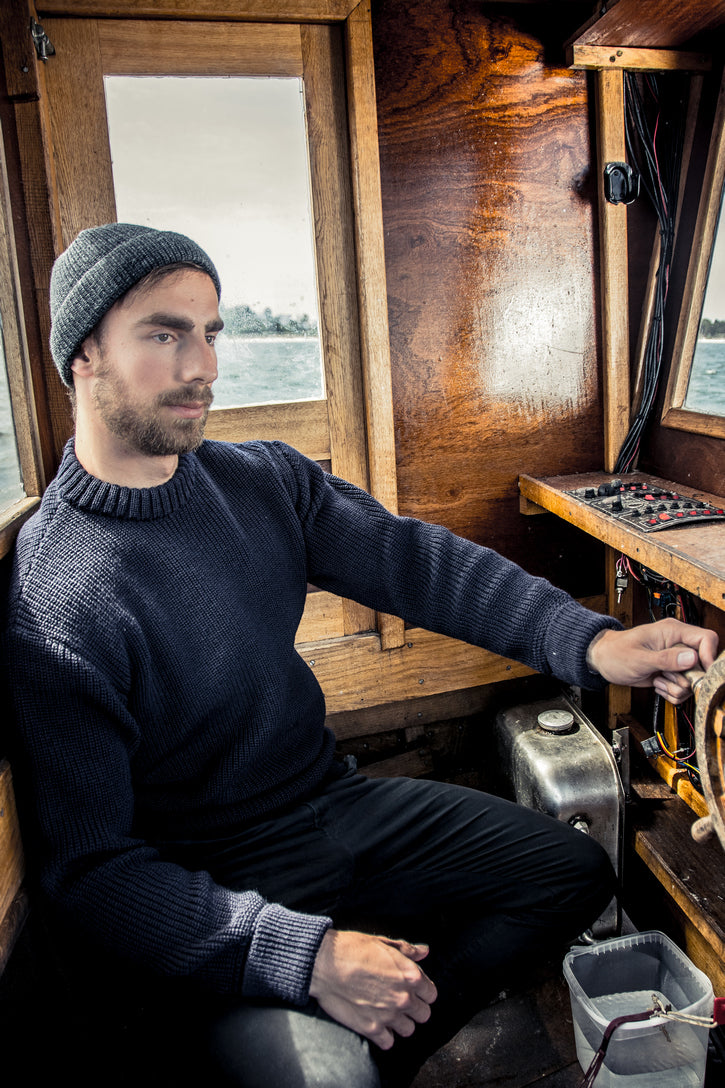 Mann in dunkelblauem Rundhals-Troyer aus 100% Schurwolle von Blauer Peter sitzt entspannt im Holzboot am Steuerrad