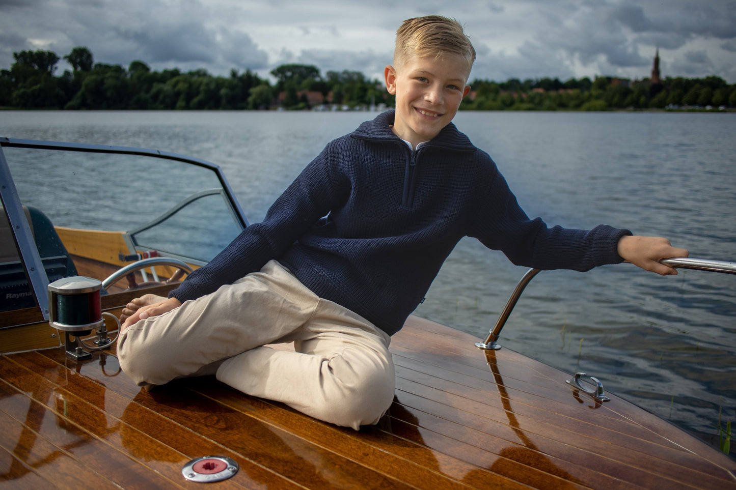 Junge im marineblauen Kinder-Troyer aus 100% Schurwolle sitzt entspannt auf einem Bootsdeck am Wasser