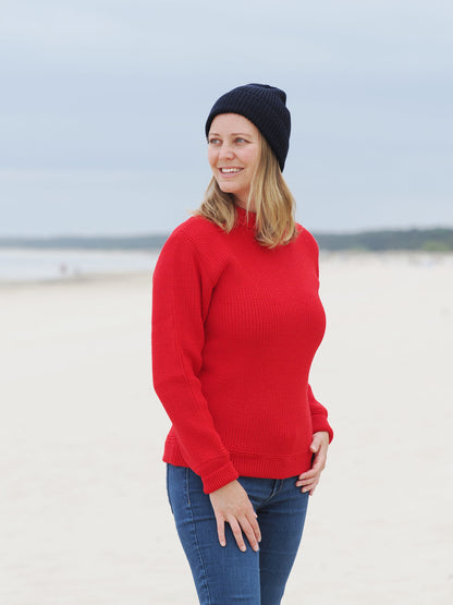 Frau steht in rotem Rundhals-Troyer und Mütze am Strand