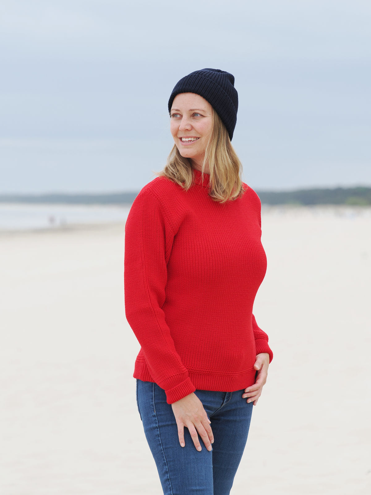 Frau steht in rotem Rundhals-Troyer und Mütze am Strand