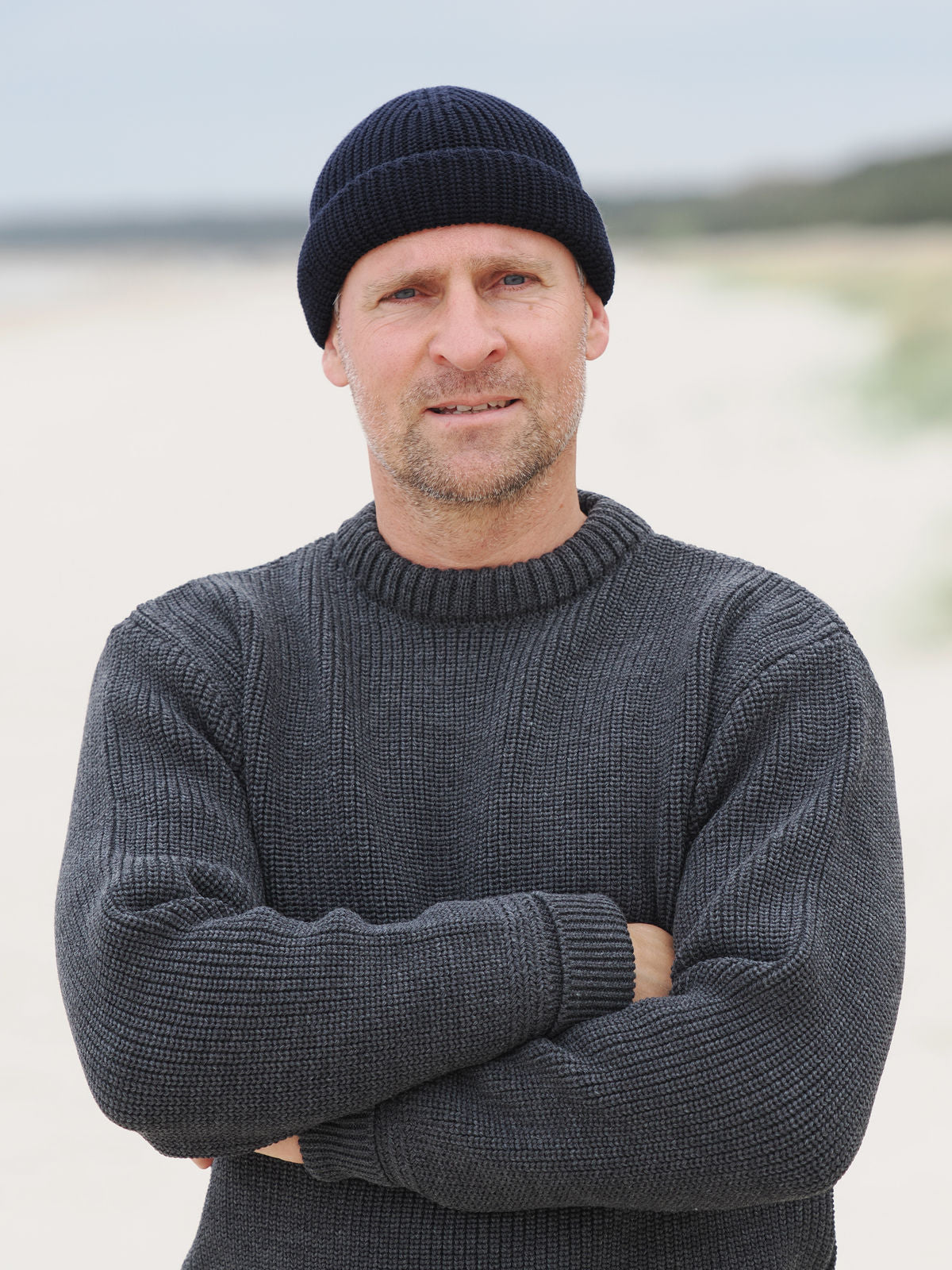 Mann steht mit verschränkten Armen im Rundhals-Troyer und Beanie am Strand