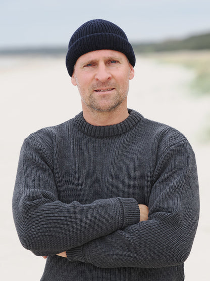Mann steht mit verschränkten Armen im Rundhals-Troyer und Beanie am Strand