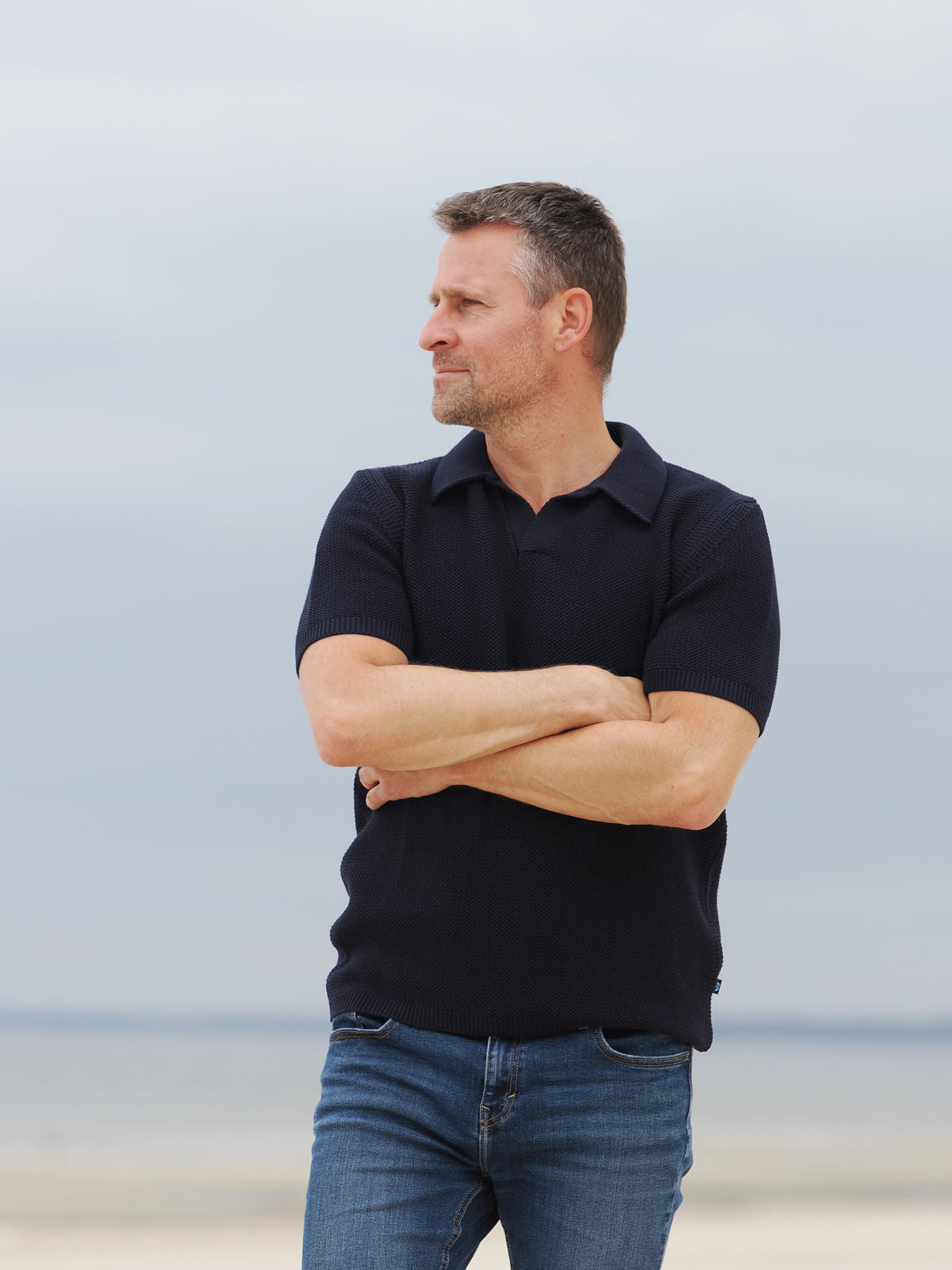 Mann im Strickpolo von Blauer Peter mit verschränkten Armen am Meer