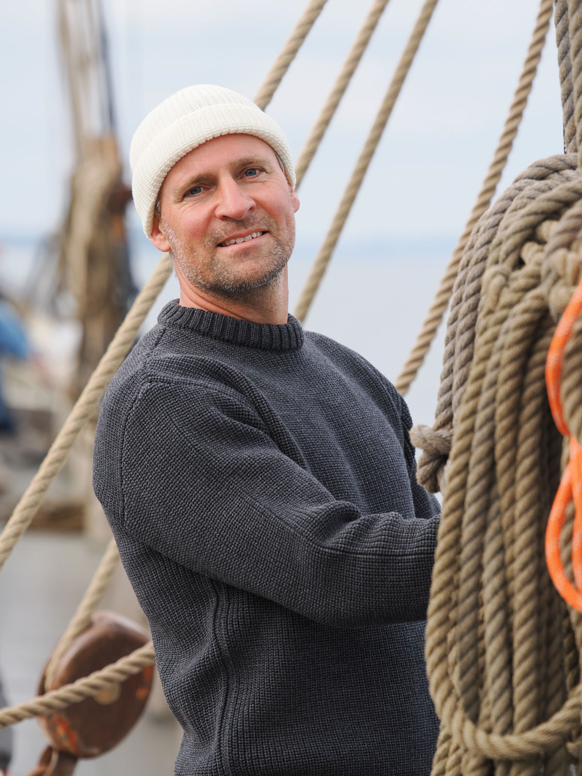 Mann steht in anthrazitem Rundhalstroyer und weißer Mütze in der Takelage eines Segelschiffs