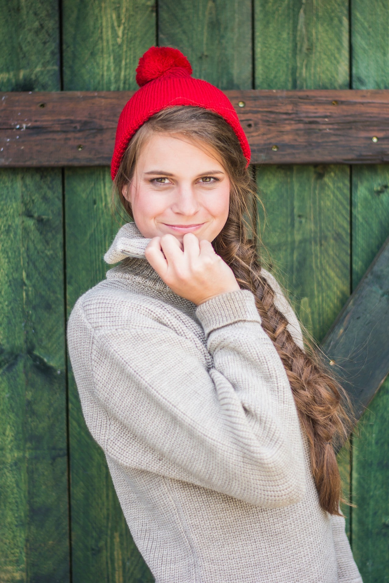 Junge Frau im beigefarbenen Schurwoll-Troyer und roter Strickmütze von Blauer Peter vor rustikalem Holzhintergrund
